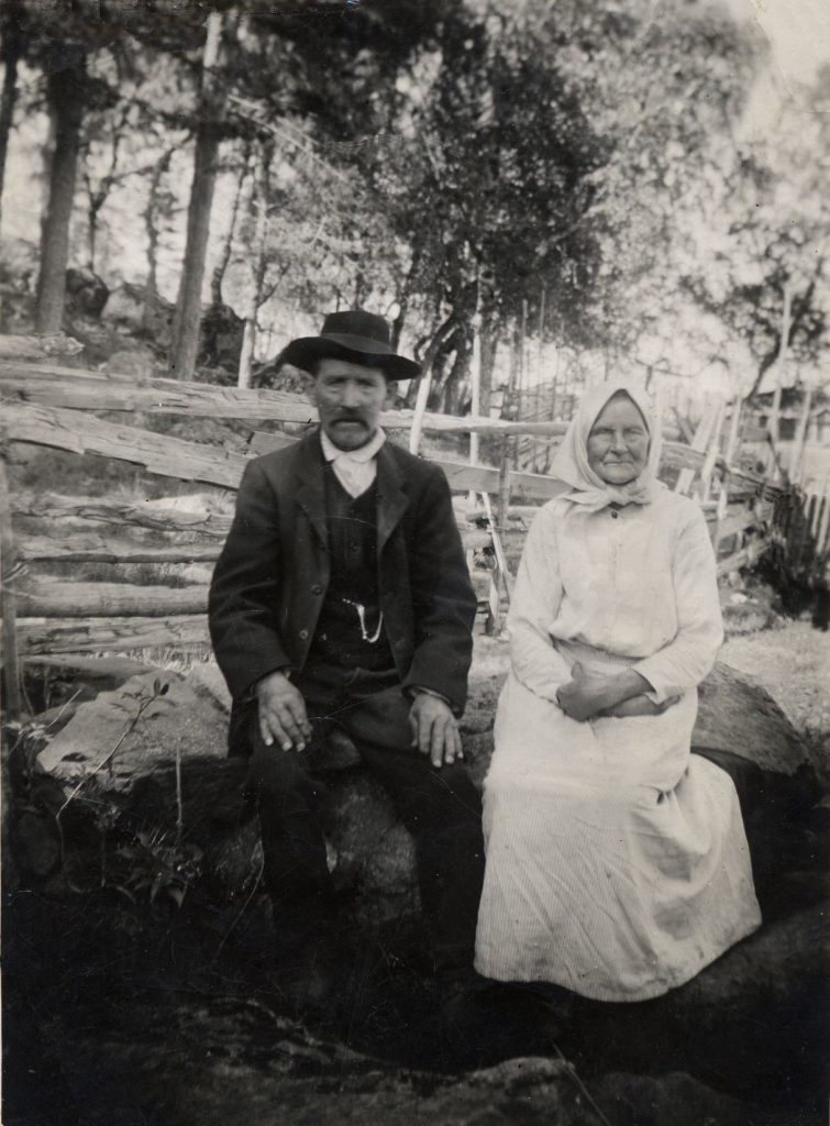 Historiallinen mustavalkoinen kuva, jossa Karin isoisän vanhemmat istuvat kiven päällä puuaita takanaan.