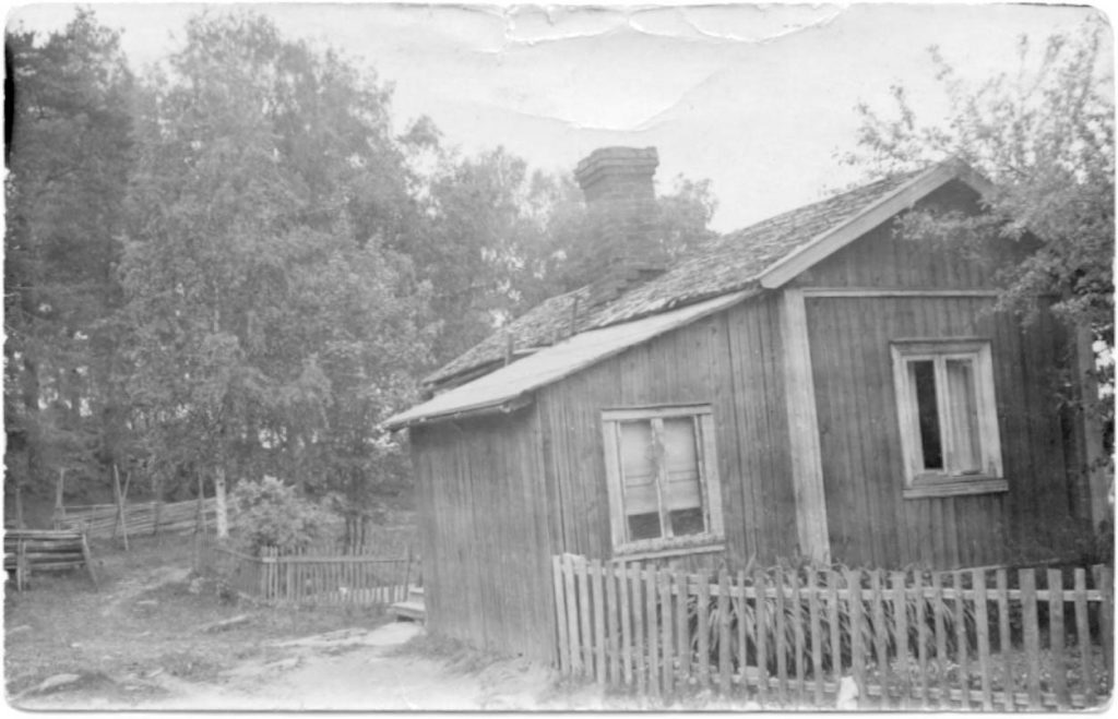 Historiallinen mustavalkoinen kuva, jossa on vanha puinen mäkitupa ja aitaa.