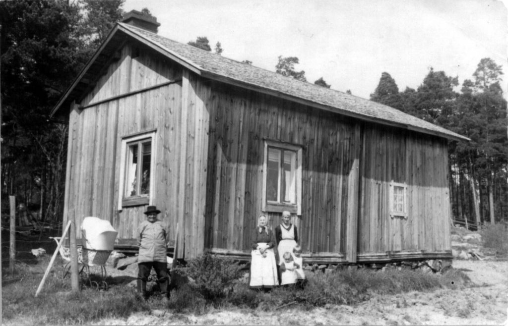 Historiallinen mustavalkoinen kuva, jossa on vanha puutalo ja sen edustalla 5-henkinen perhe.