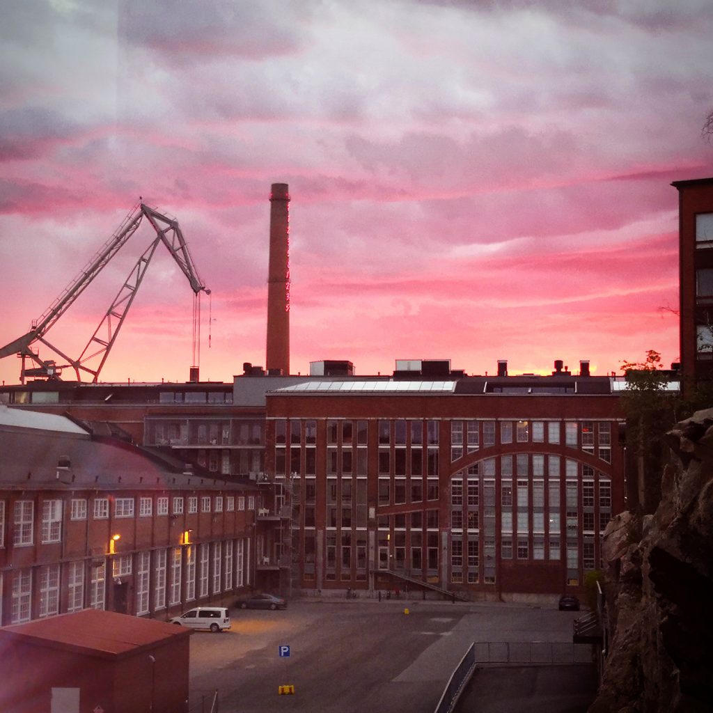 Punatiilisiä rakennuksia vasten vaaleanpunaista taivasta, jossa kohoaa voimalaitoksen piippu ja Turun Telakkarannan nosturi. 