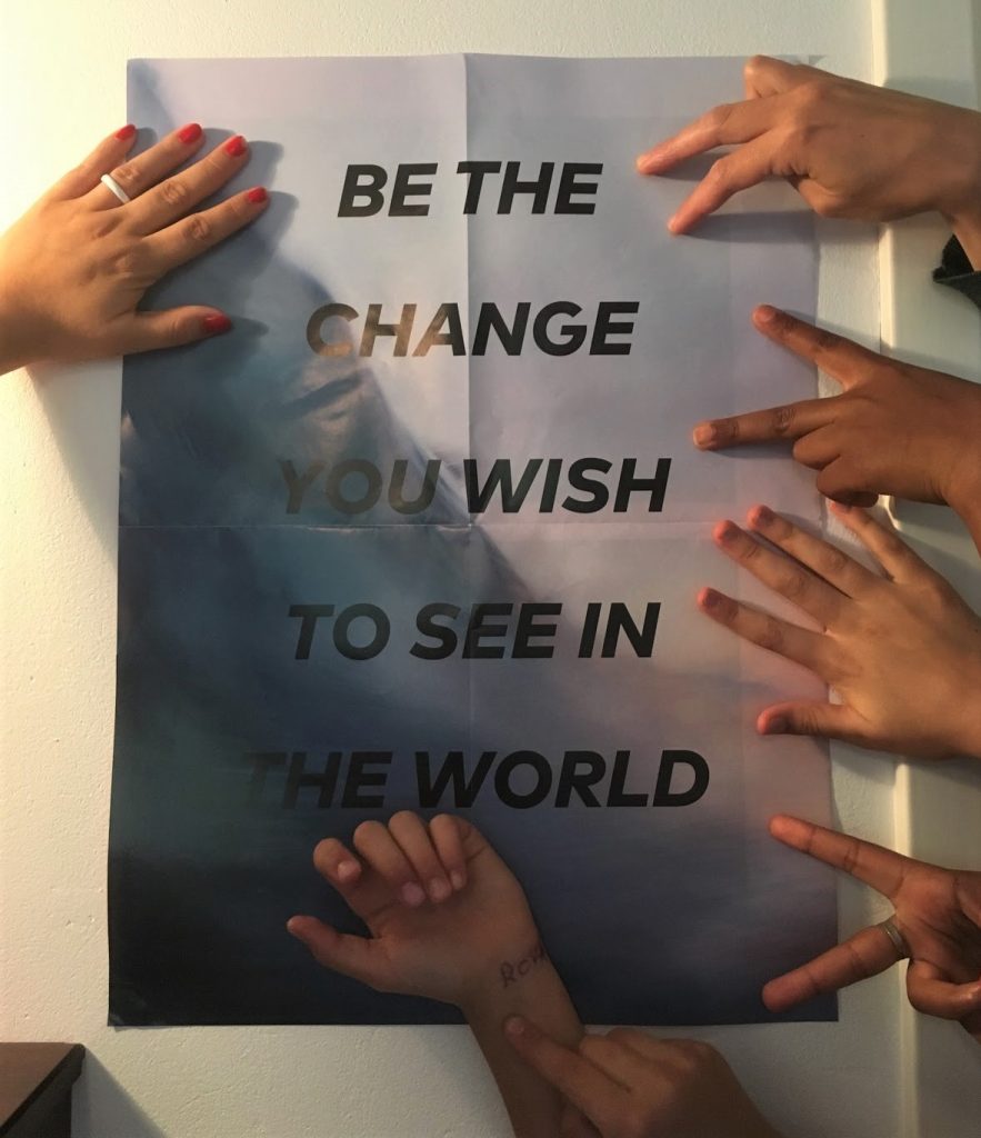 Eri-ikäisten tyttöjen käsiä posterin edessä, jossa lukee 'be the change you wish to see in the world'.