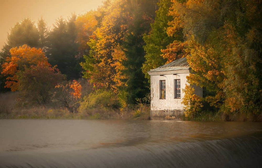 A tiny house stands on a Aura River bank where the Halinen Dam starts to fall and the autumn leaves are colouring the trees around the house.
