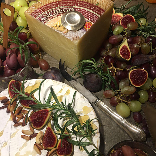 Juustopöytä, jossa Manchego ja Brie-juustoja. Tarjoilu on koristeltu runsaalla määrällä viikunoita, viinirypäleitä, oliiveja, siemeniä, hunajaa ja rosmariinin oksia.