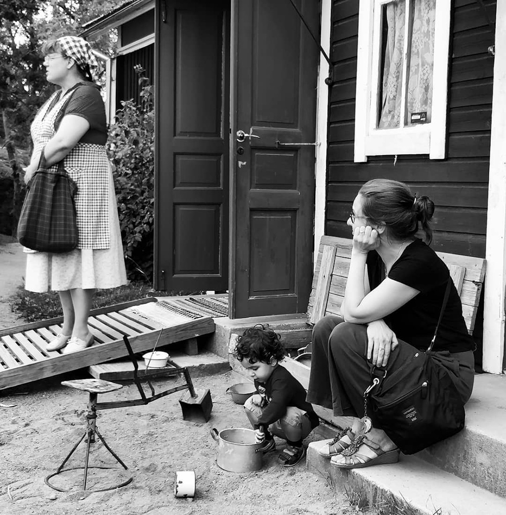 Mustavalkoinen kuva Kuralan kylämäen historiallisen ympäristön maalaistalon edestä. Nainen kuuntelee keskittyneesti oppaan kertomusta Kuralasta ja lapsi leikkii talon portailla.