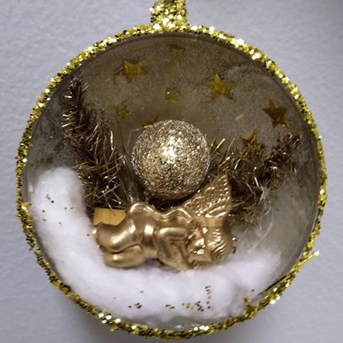 Kullanvärinen joulupallo, joka on halkaistu kahtia. Pallon sisään on rakennettu miniatyyrimaailma, jossa kultainen enkeli makaa pumpulilla kultaisen kimalluksen hohteessa. Kuvan on ottanut Pääkaupunkiseudun kierrätyskeskus.
