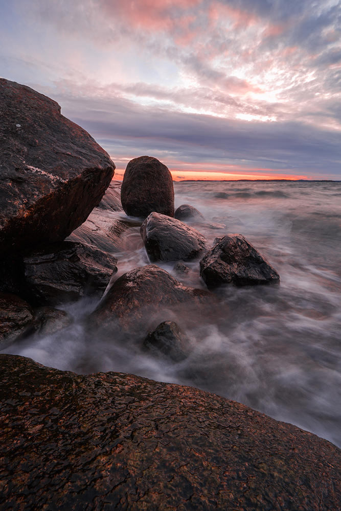 Kivikkoon osuu meren aallot Ruissalon saaren rannalla Turussa. Taustalla kaunis punertava auringonlasku.