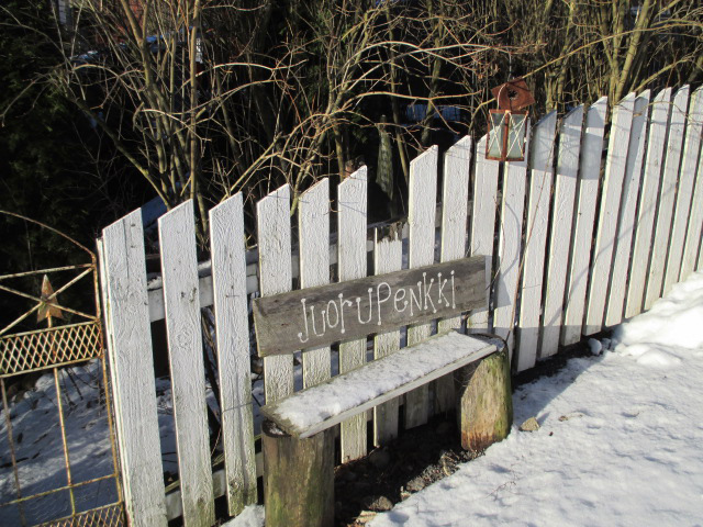 A small wooden bench and a sign behind it on a white fence saying "Juorupenkki" which means "Gossip bench" in the neighbourhood of Nummi in Turku.