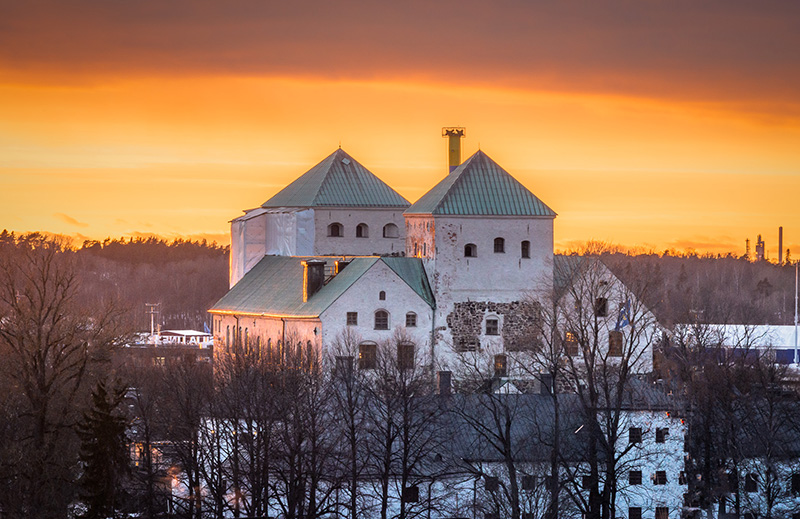Turun linna kohoaa kivisenä oranssia taivasta vasten lehdettömien puiden seasta Linnanfältin kaupunginosassa Turussa. 
