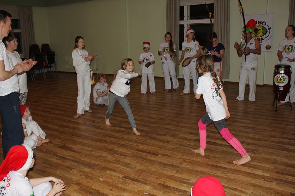 Iloisia ja nuoria capoeiran harrastajia viettämässä seuran pikkujouluja. Kaksi tyttöä ovat ringin eli rodan keskellä pelaamassa capoeirapeliä.