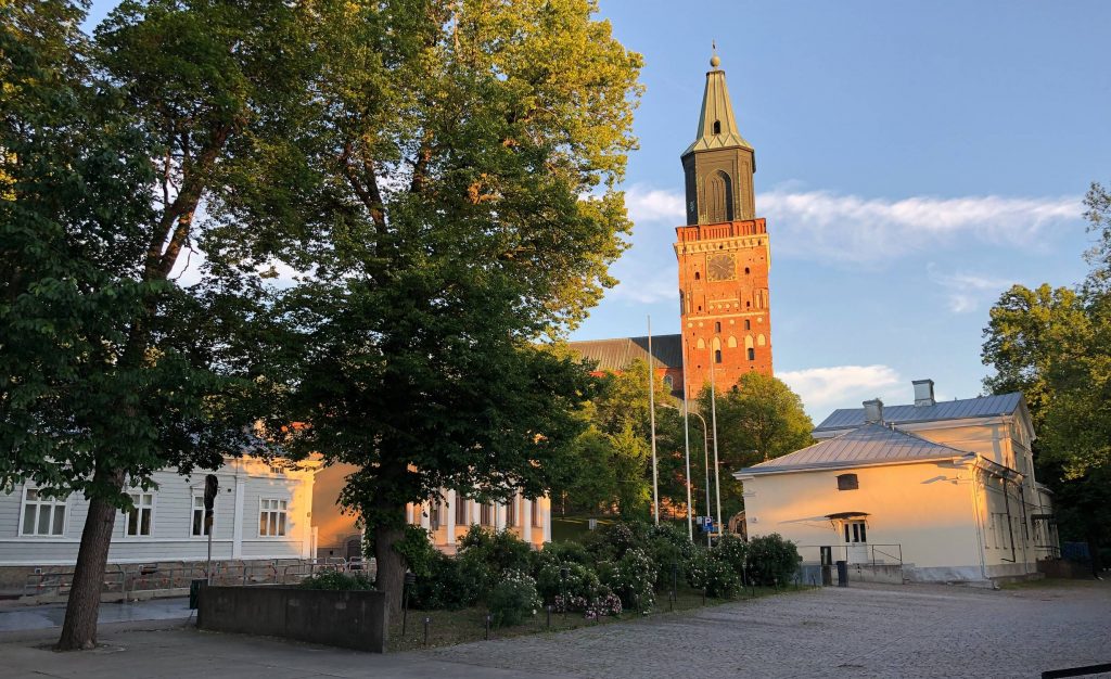 Turun Tuomiokirkko ilta-auringon valossa sinistä taivasta vasten ja muutama historiallinen rakennus, puita ja pensaita sen edustalla.