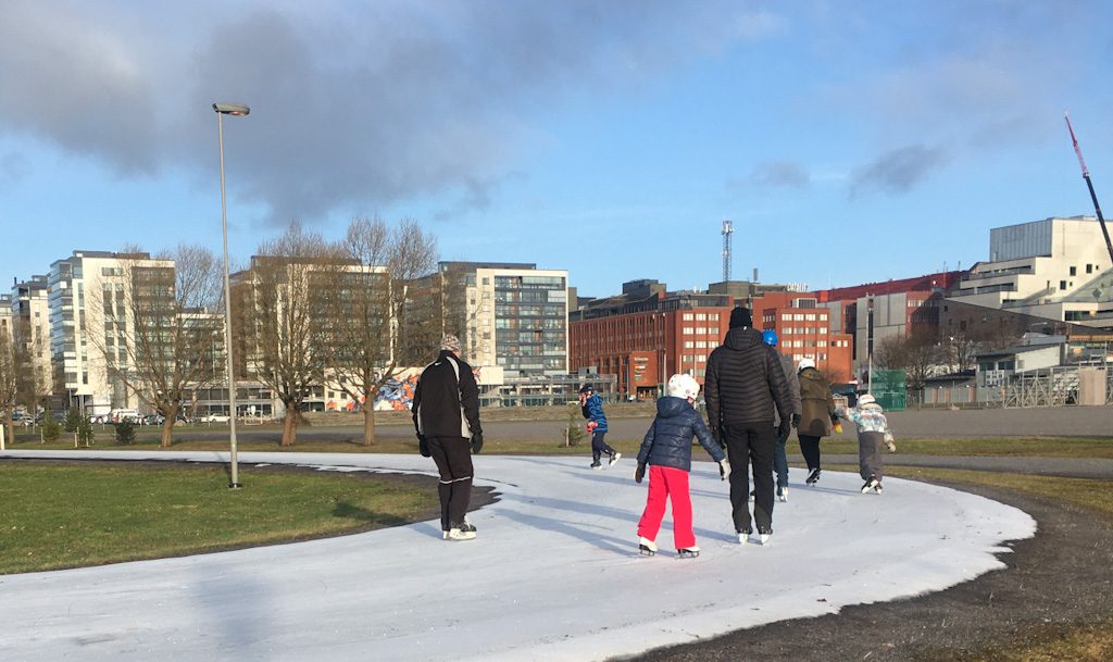 The iceskating rink in Kupittaa has a few adults and children iceskating on it on a sunny day. On the background there are modern apartment blocks.