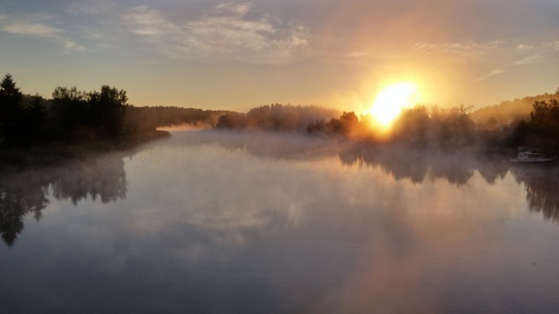 Kuusistonsalmella Kaarinassa elokuun aamuaurinko valaisee usvan läpi ja maisema heijastuu peilityynestä merestä.
