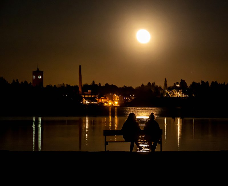 Kuutamo on värjännyt littoistenjärven syvän kultaisella sävyllä. Kaksi ihmistä istuu penkillä ihastellen maisemaa.