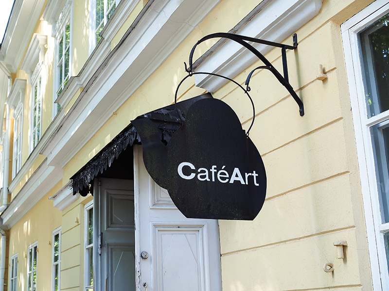 Turun Aurajoen rannassa sijaitsevan Café Artin teepannun muotoinen kyltti kahvilan oven vieressä.