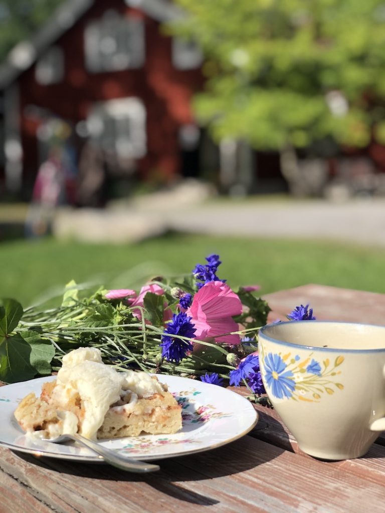 Koroisten kahvilan pöydällä omenapiiran ja kahvikuppi kesäisenä päivänä. Taustalla näkyy Koroisten kahvilarakennus. 