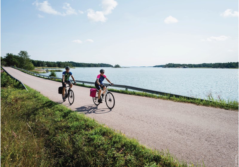 A man and a woman biking with bike bags on a concrete road by the sea on a sunny day.