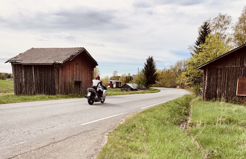 Moottoripyöräilijä asfalttitiellä ja vanhoja maaseudun puurakennuksia Hämeen Härkätiellä.