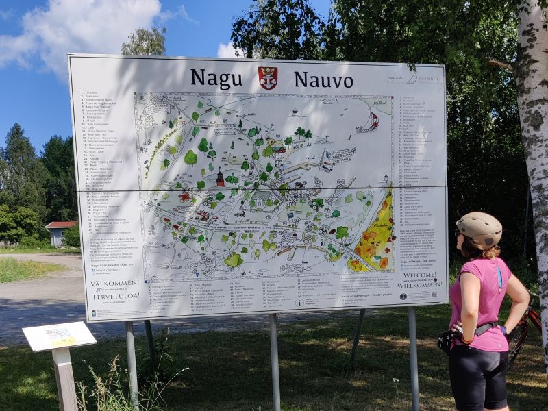 In the center of the picture is a road sign for the city of Nauvo with a beautifully drawn map of Nauvo. The author of the blog, Katerina, wearing cycling shorts, gloves, a t-shirt, helmet and sunglasses, stands to the right of the sign and examines the map. 