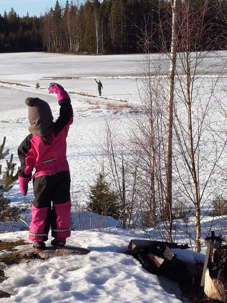 Talvinen kuva, jossa lapsi seisoo kivellä ja heiluttaa kauempana pellolla kulkevalle aikuiselle.