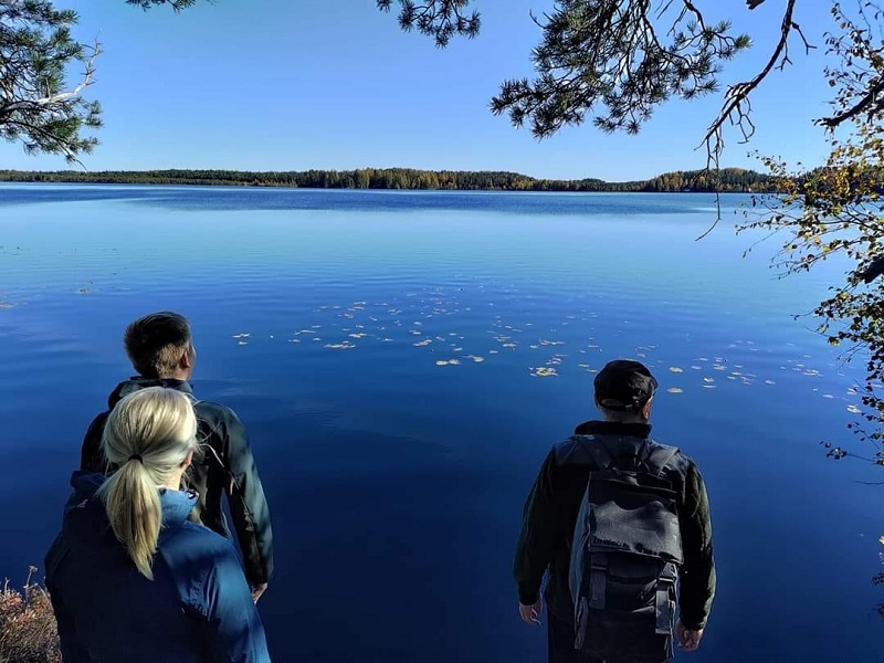Kolme retkeilijää seisoo sinivetisen järven rannalla auringonpaisteessa.