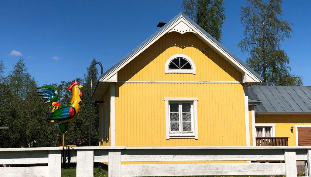 A yellow farmhouse in the sunshine, with a decorative rooster on the front fence.