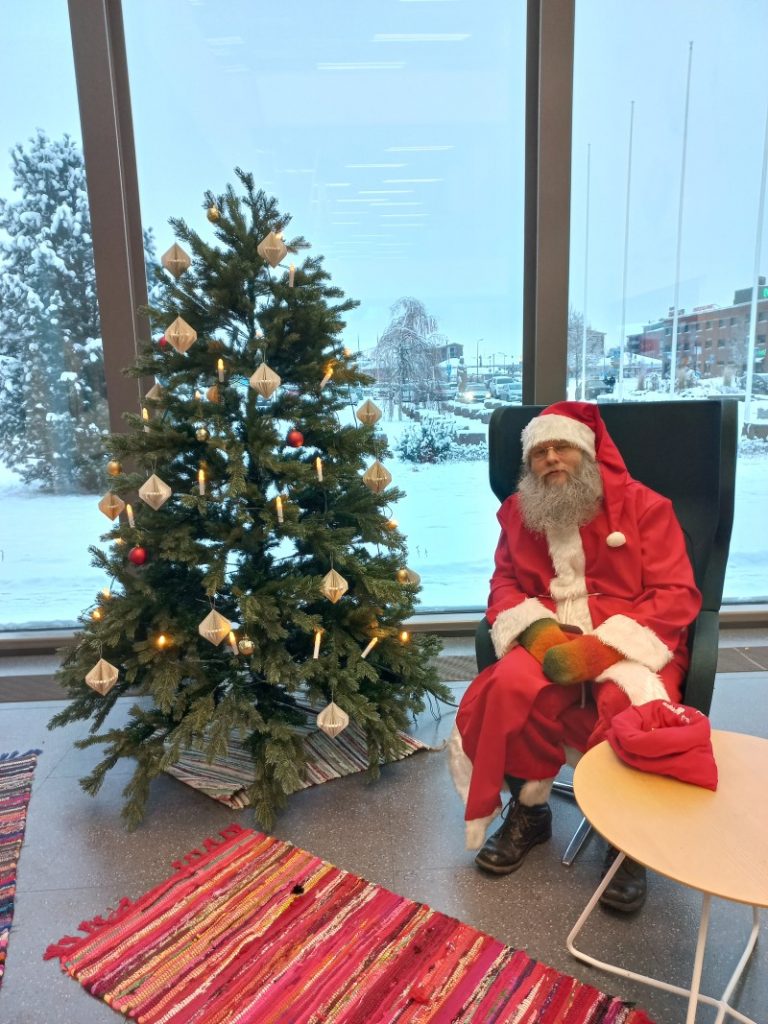 Santa Claus sits next to Christmas tree.